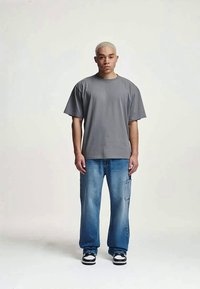 Jovem com cabelo loiro curto, vestido com uma t-shirt cinza larga, jeans azuis largos e ténis preto e branco, de pé contra um fundo simples.