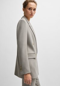 Lys grå skræddersyet blazer med spidse lapeller, dobbeltbrystet design, sidelommer og knapper i guldtoner. Glat stofstruktur.