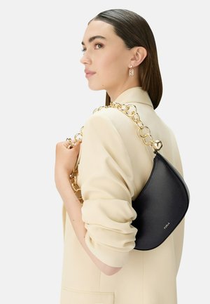Czarna skórzana torba na ramię o zakrzywionym kształcie, z złotym łańcuchem jako paskiem oraz złotym logo jako akcentem. Nosi na jasnym żakiecie.