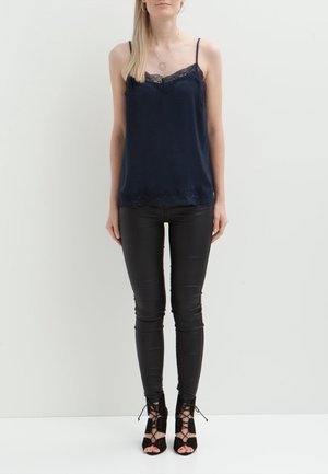 Donna che indossa una canottiera in pizzo blu navy, pantaloni skinny in pelle nera, sandali con tacco alto neri allacciati e una collana d'oro.