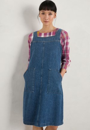 Denim overall dress with square neckline, two front pockets, and a relaxed fit. Worn over a pink plaid long-sleeve shirt.