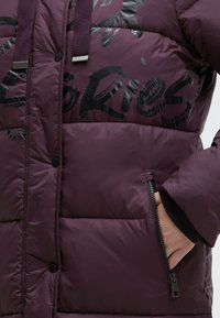 Puffer jacket in deep purple fabric, featuring a glossy black graphic design, zippered pocket, and ribbed cuffs.
