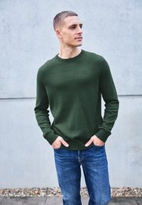 Pull en tricot vert à manches longues, encolure ronde et petit logo. Associé à un jean en denim bleu, sur un fond gris.