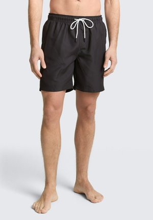 Mężczyzna w czarnych kąpielówkach z białym sznurkiem, stojący boso na jednolitym jasnym tle.