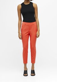 Zwarte geribbelde mouwloze top, oranje op maat gemaakte broek en zwarte instapsandalen. De broek heeft een taps toelopende pasvorm met zijzakken en een soepele textuur.