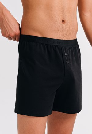 Mann trägt schwarze Baumwollboxershorts mit einem knöpfbaren Eingriff, passt den Bund an und zeigt Oberkörper und Oberschenkel gegen einen weißen Hintergrund.