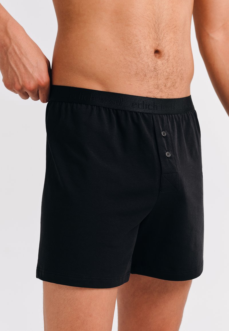 Man in zwarte katoenen boxershorts met een knoopsluiting, die de tailleband aanpast, terwijl hij zijn torso en bovenbenen toont tegen een witte achtergrond.