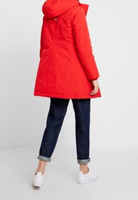 Rode geïsoleerde parka met capuchon, voorzien van een ritssluiting en knoopsluiting, gedragen over opgerolde donkerdenim broek en witte sneakers.