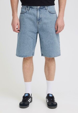 Mann iført lyseblå denimshorts, hvite crew-sokker og svarte Adidas-sneakers med hvite striper, stående på hvit bakgrunn.