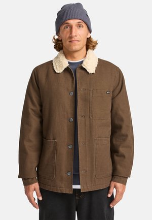 Brown cotton jacket with a plush cream collar, front pocket, and blue buttons, worn over a navy shirt and black pants.