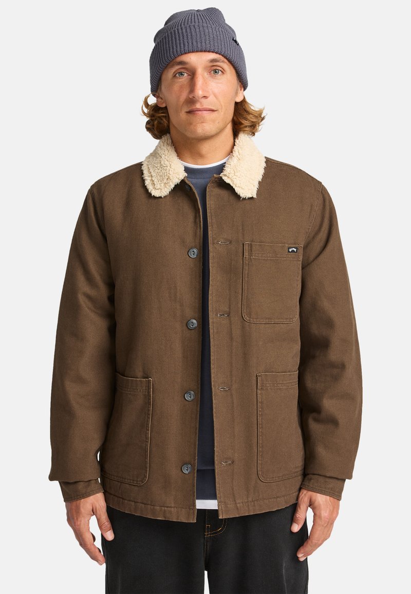 Brown cotton jacket with a plush cream collar, front pocket, and blue buttons, worn over a navy shirt and black pants.