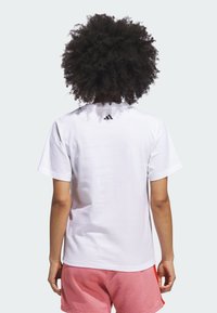 Biely bavlnený tričko s krátkymi rukávmi, na ktorom je čierne logo Adidas na hornej zadnej strane. Model má na sebe ružové šortky.