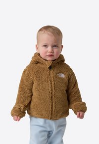 Jachetă maro din fleece cu glugă, prevăzută cu fermoar frontal și logo pe piept, purtată peste pantaloni de culoare albastru deschis. Material moale și texturat.