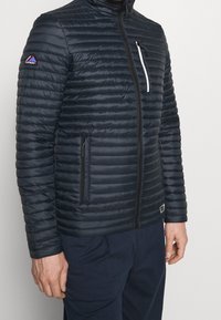 Marinblå pufferjacka med horisontell quiltad design, frontdragkedja, sidfickor och en bröstficka med vit dragkedja. Logotyp på ärmen.