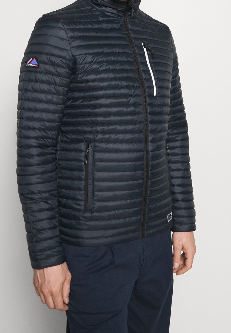 Marinblå pufferjacka med horisontell quiltad design, frontdragkedja, sidfickor och en bröstficka med vit dragkedja. Logotyp på ärmen.