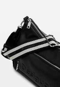 Schwarze Lederhandtasche mit strukturierter Oberfläche, ausgestattet mit einem silbernen Reißverschluss, einem verstellbaren gestreiften Tragegurt und einem geprägten Logo-Detail.