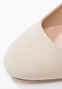 Chaussure plate pour femme en suède beige clair avec un bout arrondi et des coutures visibles le long du bord d'ouverture.