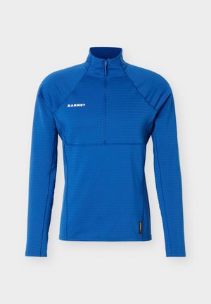 Sininen pitkähihainen kvarttivetoketjullinen urheilupaita, jossa Mammutin logo rinnassa ja Polartec-lappu helmassa, esitetty yksivärisellä taustalla.