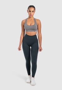 Grauer Sport-BH mit quadratischem Ausschnitt und passende, hoch taillierte schwarze Leggings. Modell trägt weiße Sneaker und steht vor einem hellgrauen Hintergrund.