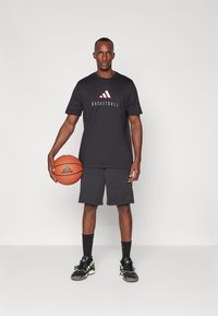 Melna sporta T-kreklā ar sarkano adidas logo, melnas šortus, melnas kedas ar zaļiem akcentiem un basketbola bumbu vienā rokā.