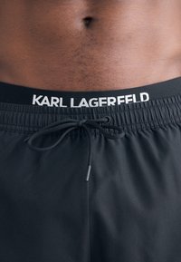 Juodos maudymosi šortai su raišteliu, ant juosmens puikuojasi kontrastingas baltas "KARL LAGERFELD" logotipas. Sklandus audinio tekstūra.