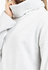 Sudadera gris claro con un cuello alto y doblado, mangas anchas y un pequeño logo en el pecho. El material parece suave y texturizado.