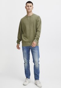 Olive green sweatshirt with a crew neck and ribbed cuffs, paired with light blue denim jeans and white sneakers. Simple design, no patterns.