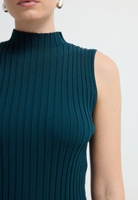 Top senza maniche a costine verde acqua con collo alto. Il tessuto presenta righe verticali e una sottile texture, evidenziando la forma.