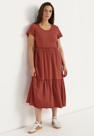 Kvinde står op ad en hvid væg iført en rustfarvet midi-kjole med korte ærmer og hvide sneakers med brune striber.