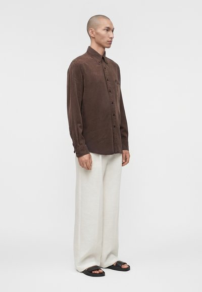 Ασιάτης άνδρας στέκεται, φοράει ένα καφέ πουκάμισο με κουμπιά, λευκό άνετο παντελόνι και μαύρα σανδάλια με σταυρωτό λουρί, σε απλό λευκό φόντο.