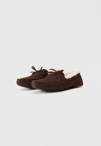 Par de zapatillas de mocasín de ante marrón oscuro con forro de piel de color crema y lazos decorativos en la parte superior, sobre un fondo liso.