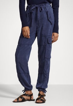 Marineblauwe cargobroek met taillekoord en enkelties, gedragen met zwarte sandaaltjes met bandjes en een donker shirt met lange mouwen.