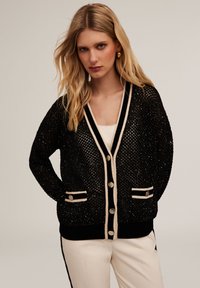 Cardigan nero lavorato a maglia con dettagli metallici, bordi beige a contrasto, due tasche frontali e chiusure con bottoni dorati. Vestibilità casual.