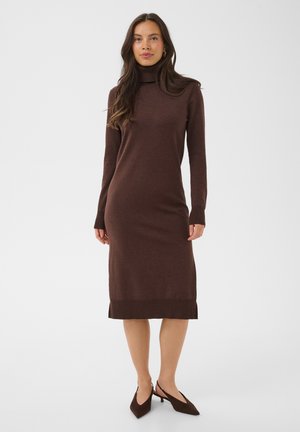Femme debout, portant une robe pull marron à manches longues, col roulé, longueur genou, avec des chaussures à talons marron foncé, sur un fond uni.