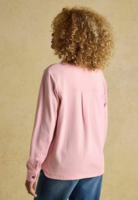 Kerge roosa pika varrukaga särk, mis on tehtud pehmest kangast, tagaküljel on plekk, nööpidega mansetid ja käisel on logoga kaunistus.