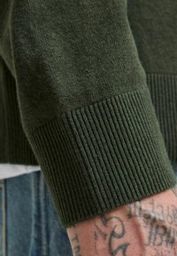 Grøn strikket trøje med detaljer som ribbet manchet og blød tekstur, der viser et close-up af en hånd med synlige tatoveringer.