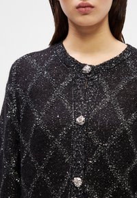 Black knitted cardigan featuring a diamond pattern with silver accents. It has round buttons and a textured neckline.