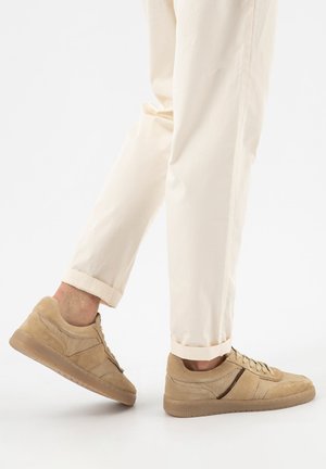 Person trägt beigen Wildledersneaker und cremefarbene hochgekrempelte Hose, ein Fuß leicht angehoben vor weißem Hintergrund.