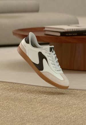 Witte en beige sneaker met zwarte details en rubberen zool zwevend boven een beige tapijt, houten salontafel en gestapelde boeken op de achtergrond.