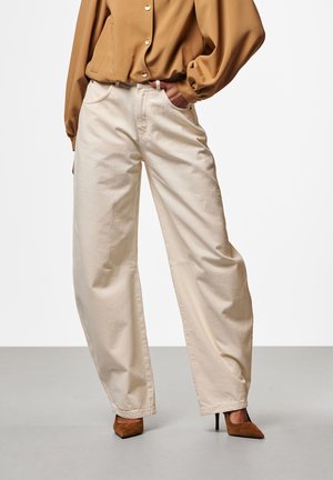 Wijde off-white broek gedragen met een bruine blouse met lange mouwen en knopen en bruine puntige hoge hakken, hand in de zak.