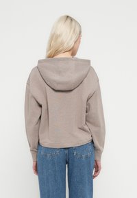 Oseba s svetlimi lasmi, oblečena v taupe kapucar in modre kavbojke, stoji z zadnjico obrnjeno proti kameri na enobarvnem belem ozadju.