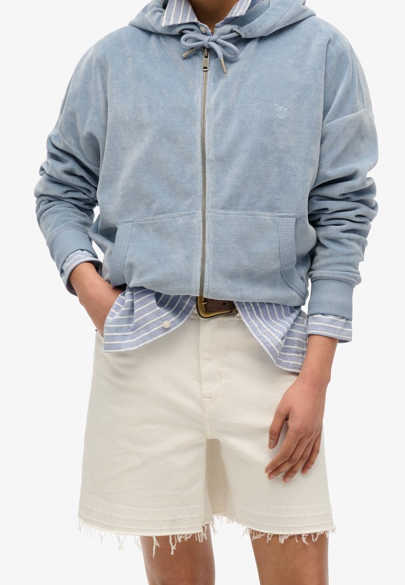 Personne portant un sweat à capuche zippé bleu clair sur une chemise rayée bleue et blanche rentrée dans un short écru effiloché avec une ceinture marron.