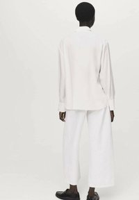 White long-sleeve shirt with a relaxed fit, soft fabric, and buttoned cuffs, paired with wide-leg white trousers and black shoes.