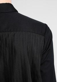 Black blouse with a classic collar, featuring a smooth fabric with vertical pleats at the back. Textured surface, simple design.