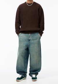 Ruskea ribbineulepaita, rento vaaleansiniset farkut ja vihreät lenkkarit valkoisilla nauhoilla, seisomassa yksiväristä valkoista taustaa vasten.