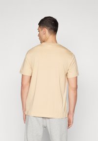 Beige Baumwoll-T-Shirt mit Rundhalsausschnitt und kurzen Ärmeln, das eine lockere Passform und minimale Nähte auf der Rückseite aufweist. Graue Hosen sind teilweise sichtbar.