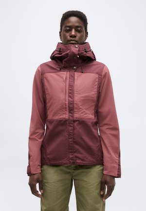 Person iført en tofarvet vinrød hættetrøje med høj krave og olivengrønne bukser, stående mod en ensfarvet lysegrå baggrund.