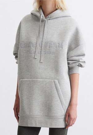 Sweat à capuche oversize gris clair avec poche kangourou et texte en relief "Marc O'Polo Est. in Stockholm,", porté avec les manches remontées.