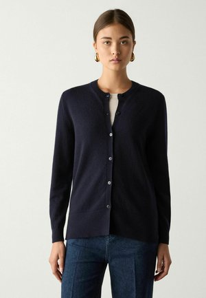 Frau trägt eine dunkelblaue, geknöpfte Strickjacke über einem hellen Oberteil, kombiniert mit blauen Jeans, frontal vor einem einfarbigen hellen Hintergrund.