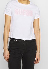 Hvid cropped t-shirt lavet af bomuld med korte ærmer, prydet med et blankt lyserødt "GUESS" logo på forsiden. Kombination med sorte jeans.